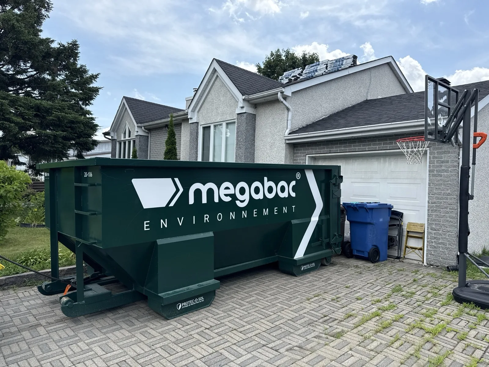 20 cubic yard Megabac dumpster delivered on a paver driveway for a roof replacement — bundles of new shingles stacked on the roof